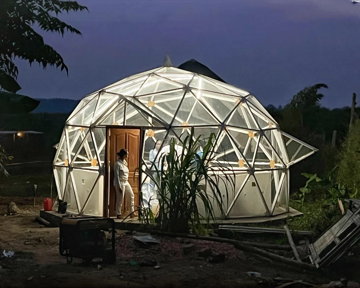 Illuminated geodesic-dome greenhouse at Bioksanland at twilight, with a person standing in the wooden doorway of the structure