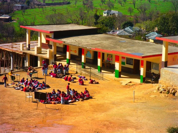 A school building and groups of students gathered in its grounds, surrounded by green hills